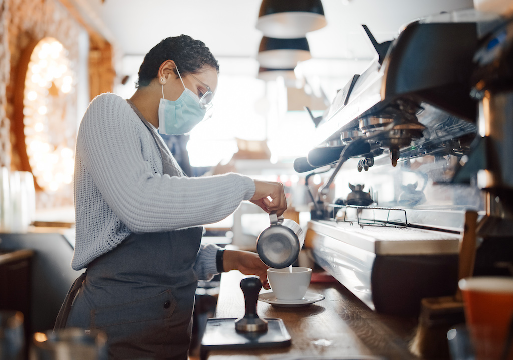 Beautiful Latin American Female Barista in Face Mask is Making a Cup of Cappuccino in Coffee Shop Bar. Social and Medical Health Restrictions Concept in a Loft-Style Cafe During Coronavirus Pandemic.