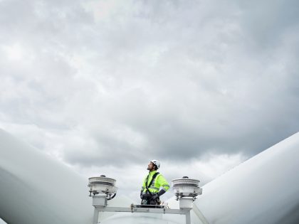 Professional rope access technician standing on roof (hub) of wind turbine between blades and antennas and looking up on the blade. Dramatic sky behind. Hands on hips