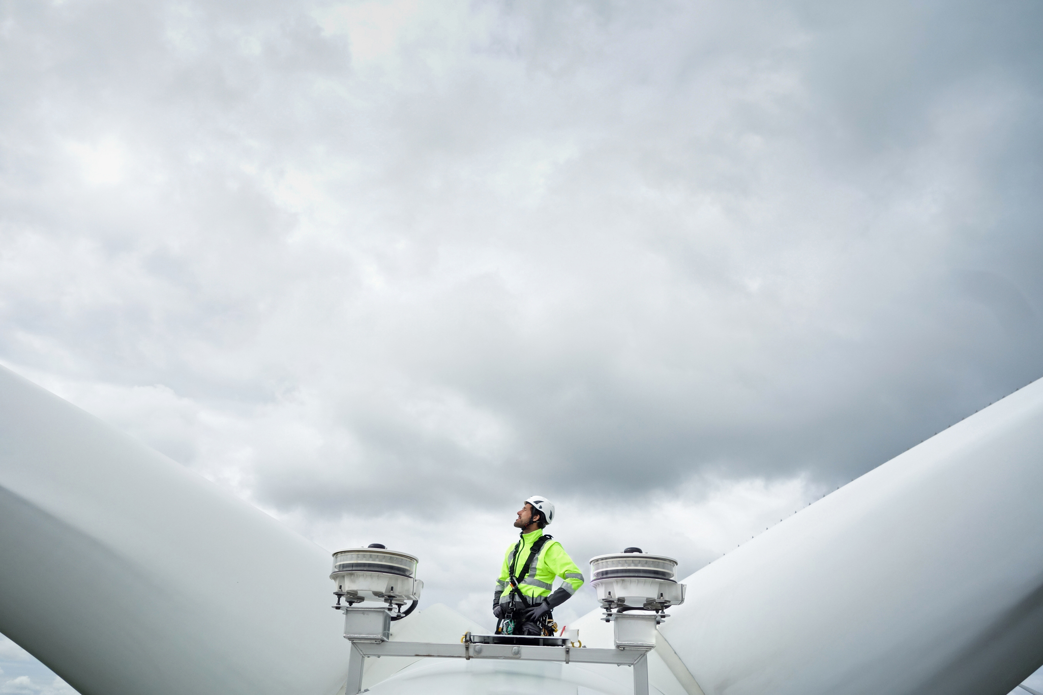 Professional rope access technician standing on roof (hub) of wind turbine between blades and antennas and looking up on the blade. Dramatic sky behind. Hands on hips