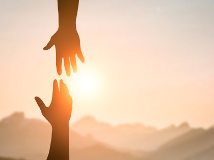 Silhouette of two people hands reaching to each other for help in sunset sky and orange sun. Friendship, teamwork, help, faith and hope concept.