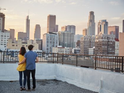 Rear View Of Couple On Rooftop Terrace Looking Out Over City Skyline At Sunset