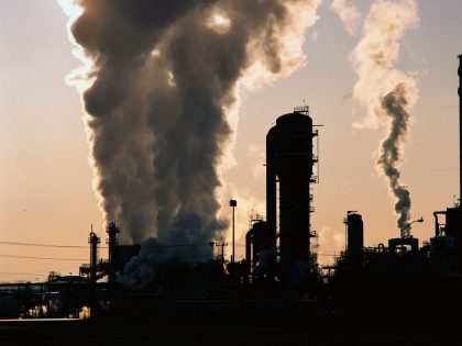 This is an image of an industrial plant pushing billowing stacks of smoke into the sky. It is a petroleum processing plant that is clearly showing its contribution of pollution to the environment. The plant is silhouetted against a somewhat grayish s