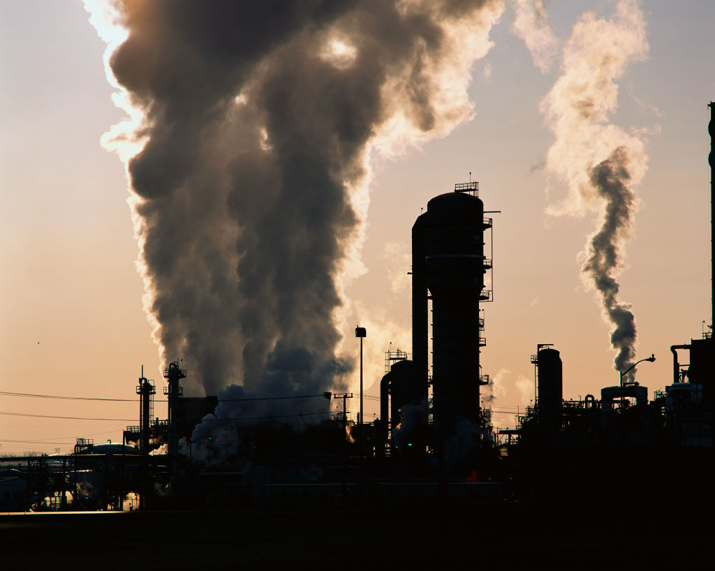 This is an image of an industrial plant pushing billowing stacks of smoke into the sky. It is a petroleum processing plant that is clearly showing its contribution of pollution to the environment. The plant is silhouetted against a somewhat grayish s