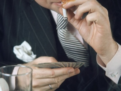 A man smokes a cigarette while playing cards, with an alcoholic drink in the foreground.
