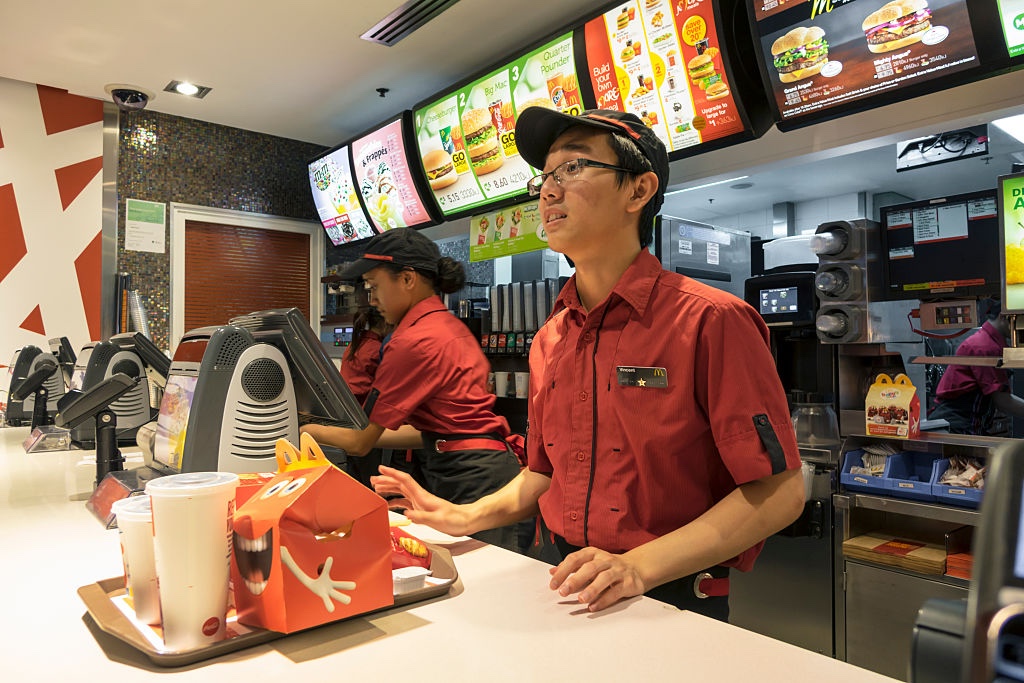 A young McDonald’s worker stands behind the counter at a McDonald’s restaurant with a tray with an order of food on it.