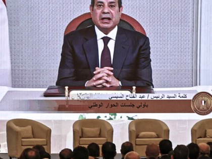 Abdel Fattah al-Sisi sitting on a chair at a table on a screen in front of a crowd.