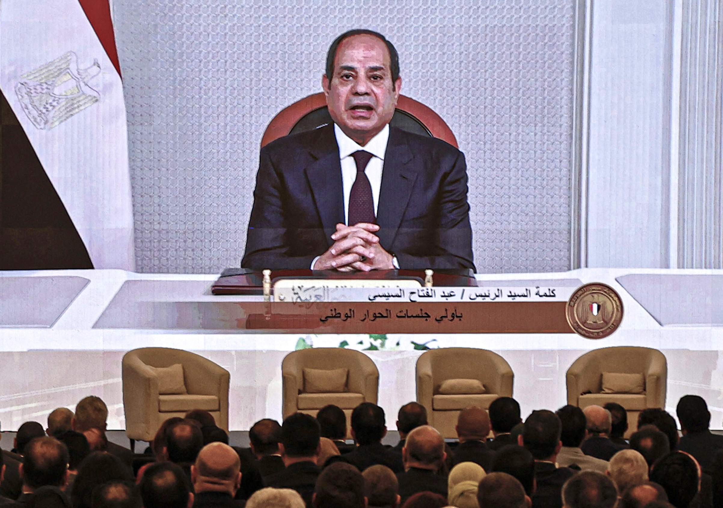 Abdel Fattah al-Sisi sitting on a chair at a table on a screen in front of a crowd.