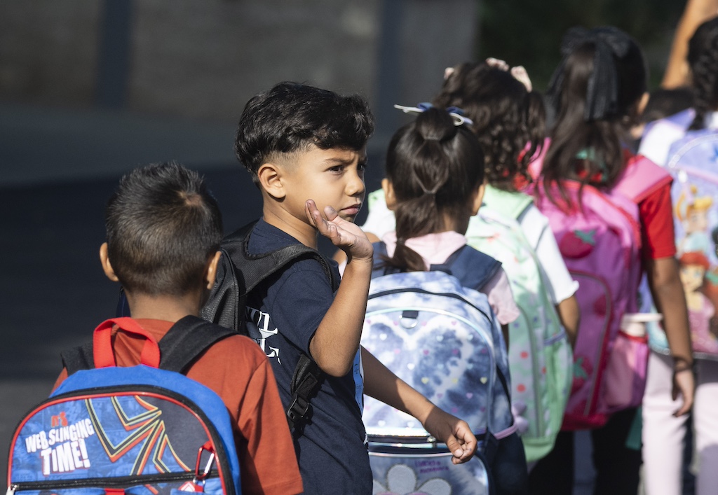 An elementary-aged student turns and waves as he walks in a line with other children.