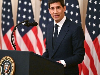 Kevin Warsh standing at a podium with a row of American flags behind him.