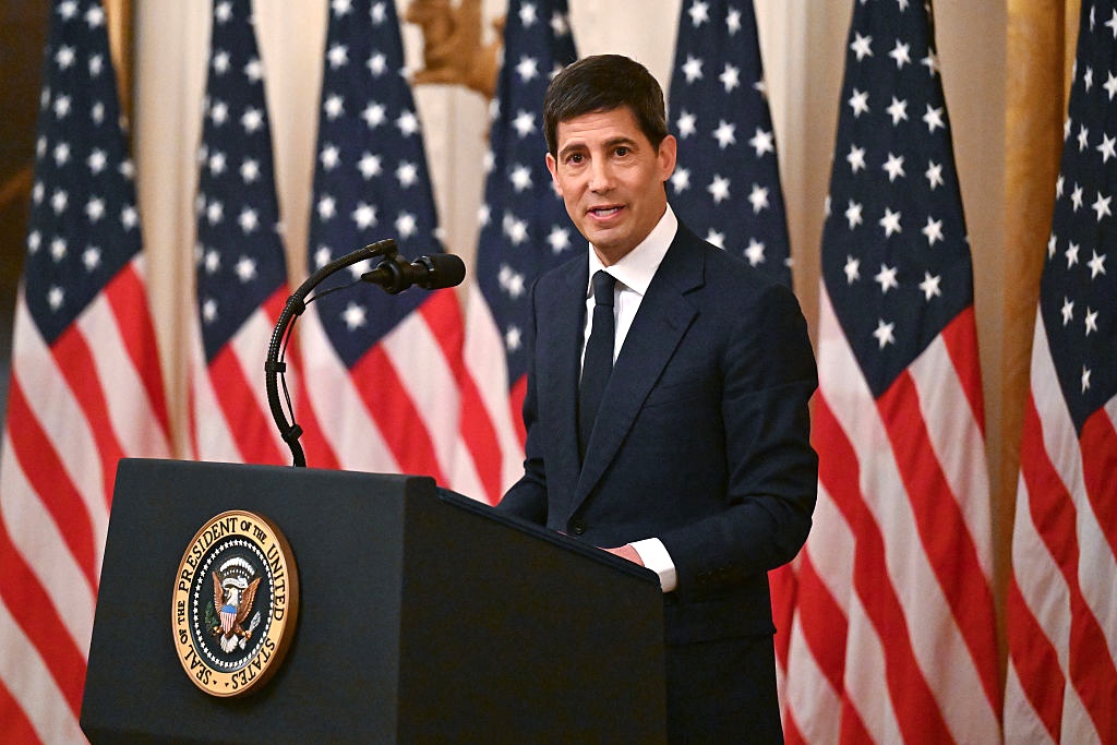 Kevin Warsh standing at a podium with a row of American flags behind him. 