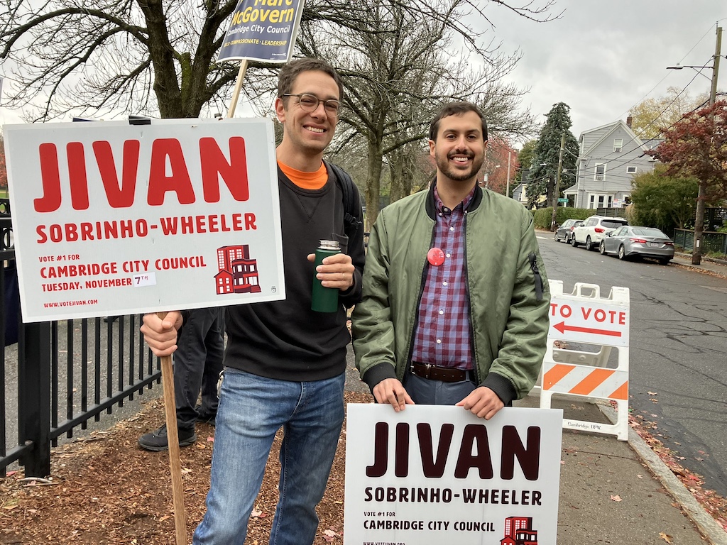 Jivan Sobrinho-Wheeler stands with a supporter, both holding campaign signs for Jivan. 