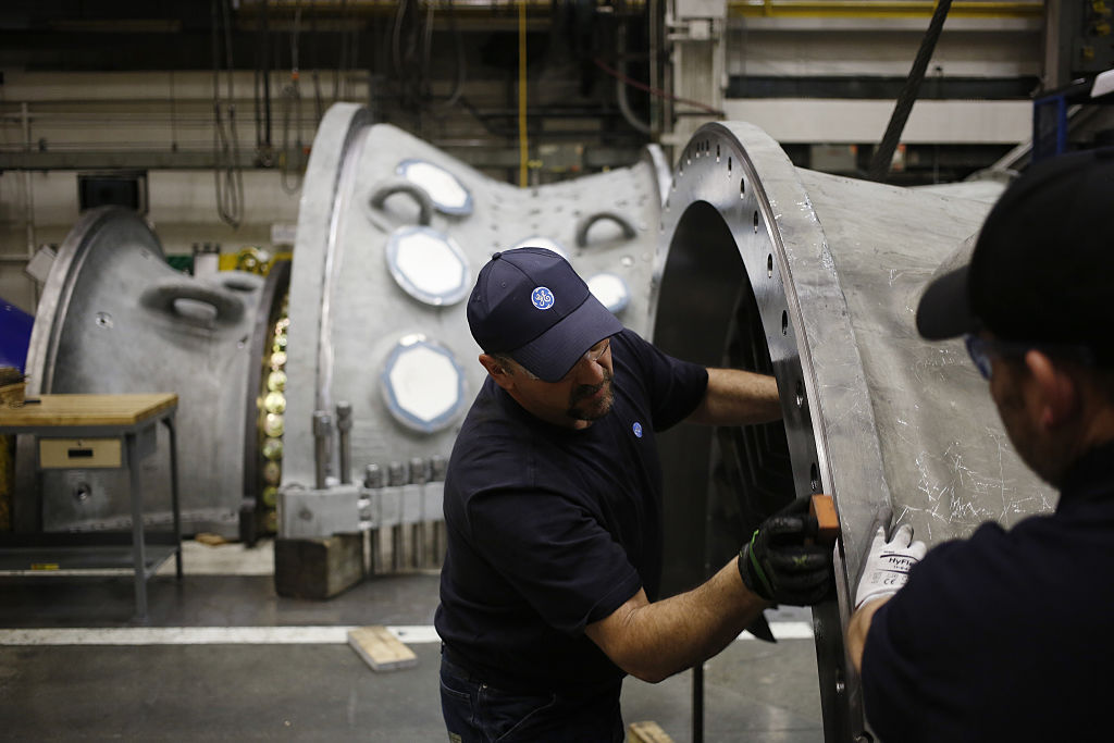 A man works on large metal machinery as another worker looks on.