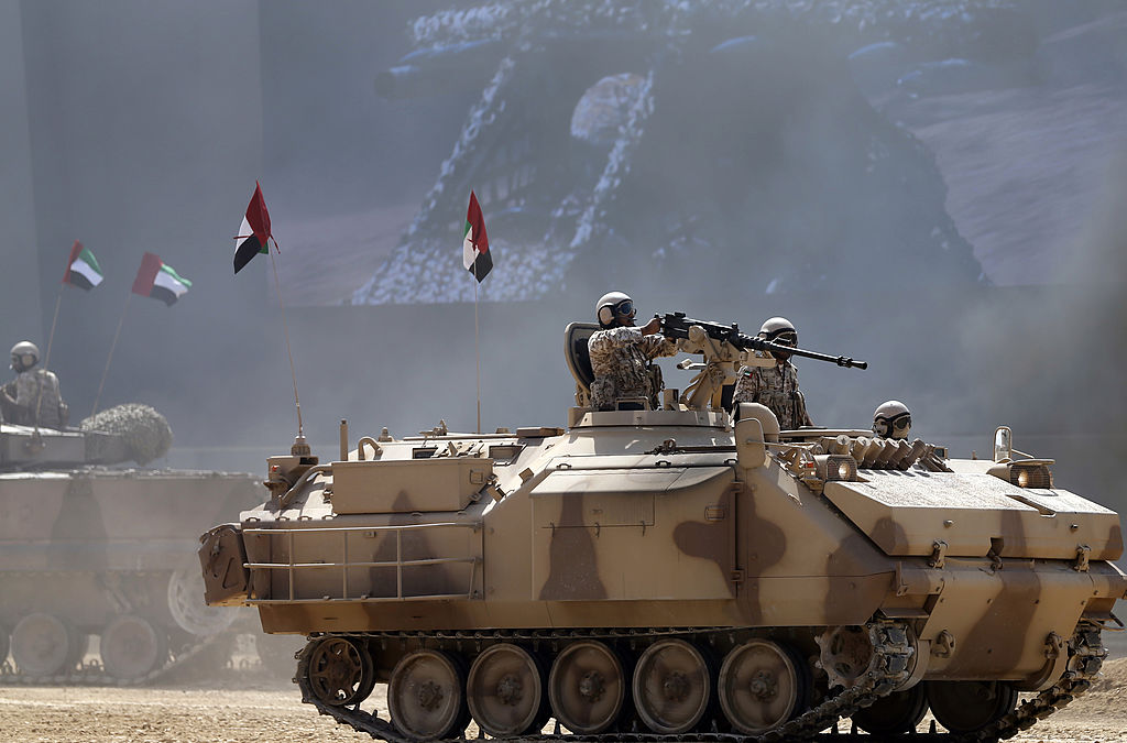 Emirati armed forces personnel in a battle tank with guns drawn and UAE flags in the background.