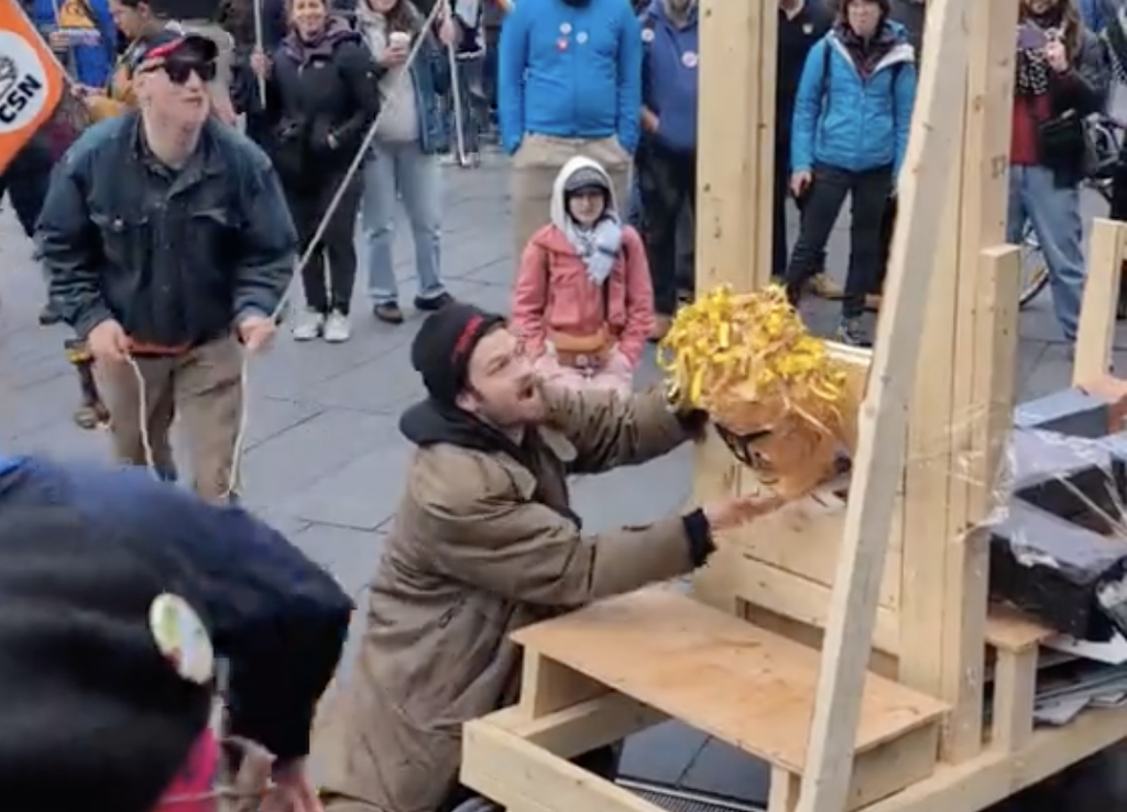 A man holds a head in a guillotine while a crowd looks on.