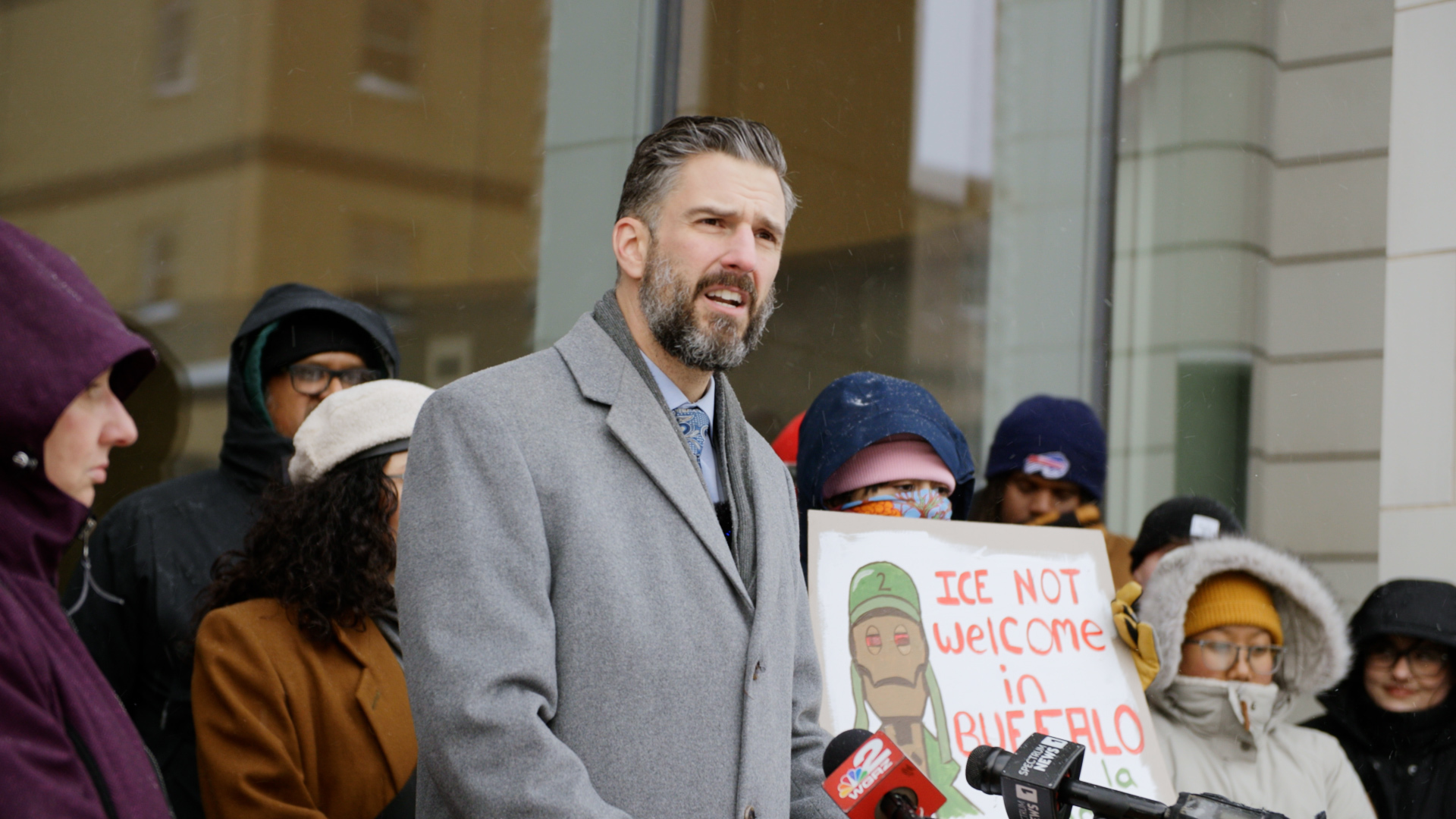 Adam Bojak speaking while a crowd surrounds him, one person holding up a sign that says, 