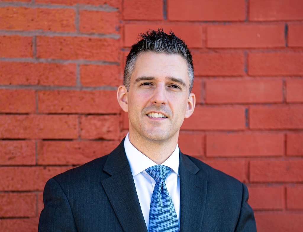 Portrait of Adam Bojak in a suit against a brick wall.