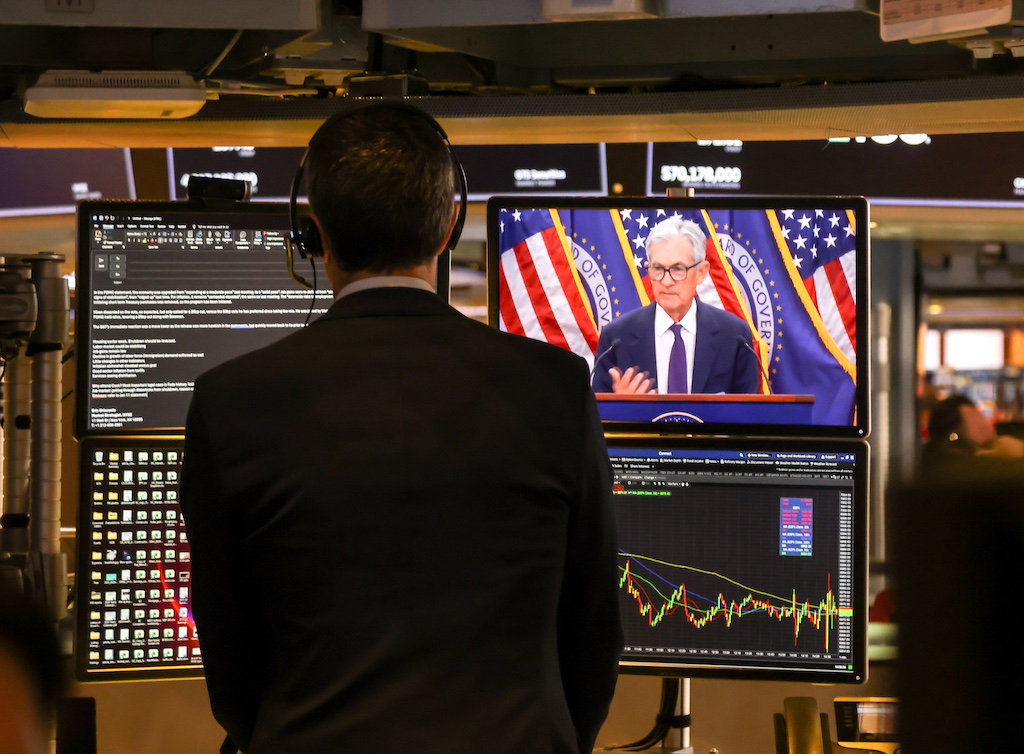 A man stands facing four TVs at the New York Stock Exchange, one showing Fed chair Jeremy Powell. 
