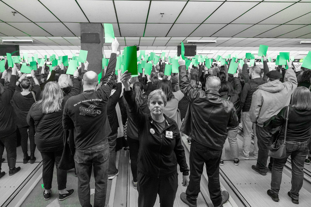 An image in black and white of a crowd of people holding up cards that are in color, green.