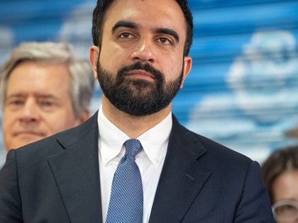 Zohran Mamdani standing in a suit with two people behind him.