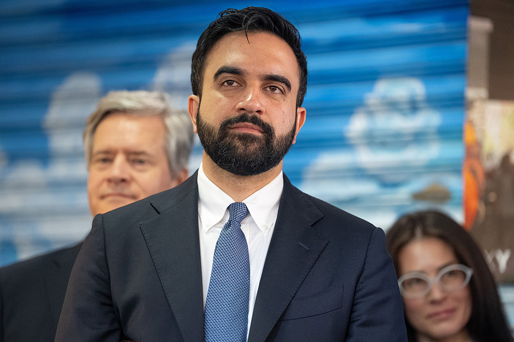 Zohran Mamdani standing in a suit with two people behind him.