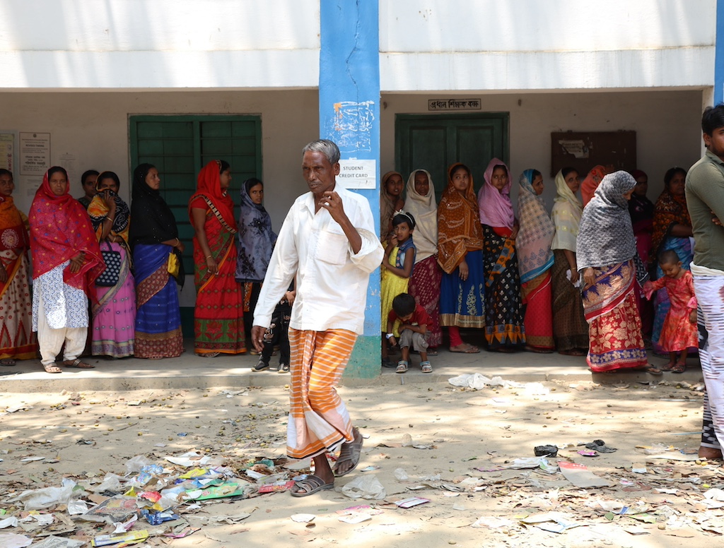 A man walks away from a line of people waiting against a wall in the shade.