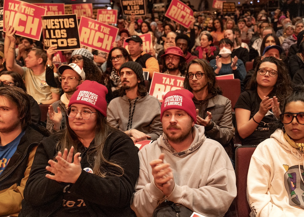A crowd of people, some holding up placards that say "Tax the Rich" and some wearing hats that say "Democratic Socialists of America."
