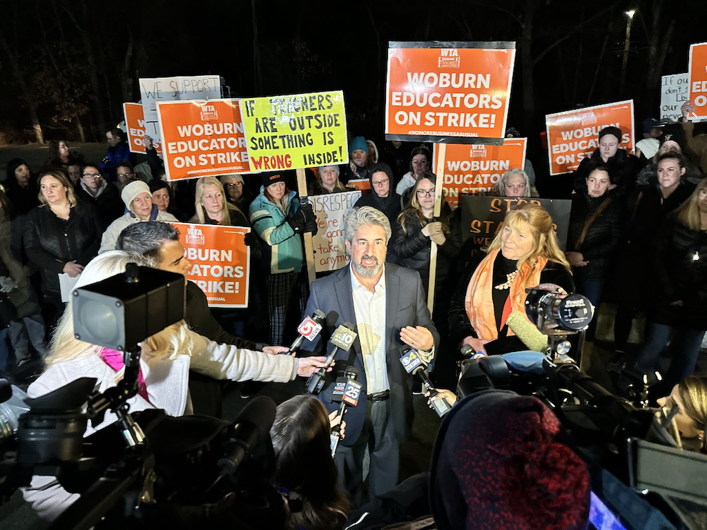 A man speaking to media with people surrounding him with picket signs.