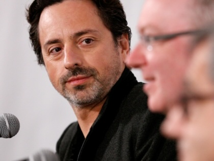Sergey Brin in front of a microphone looking on as another person sitting next to him speaks. 