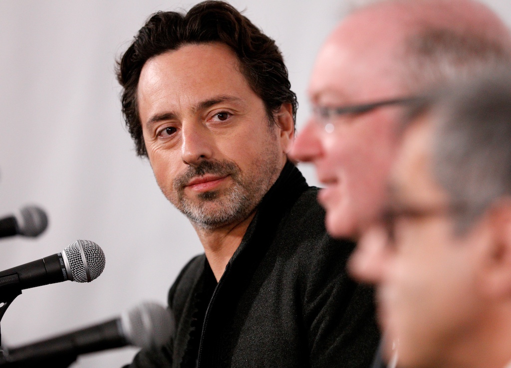 Sergey Brin in front of a microphone looking on as another person sitting next to him speaks. 
