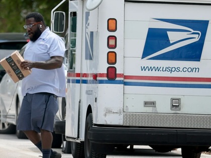 A USPS mail carrier holds an Amazon package while walking away from a parked USPS mail truck.