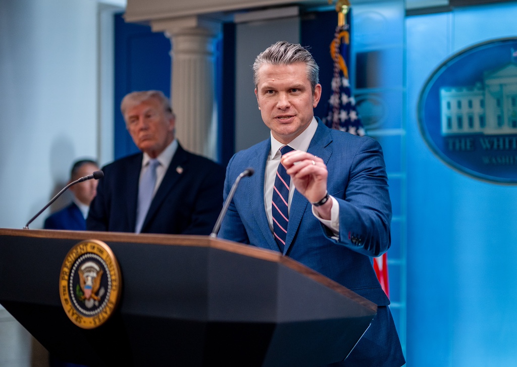 Pete Hegseth speaking at a podium with Donald Trump looking on.