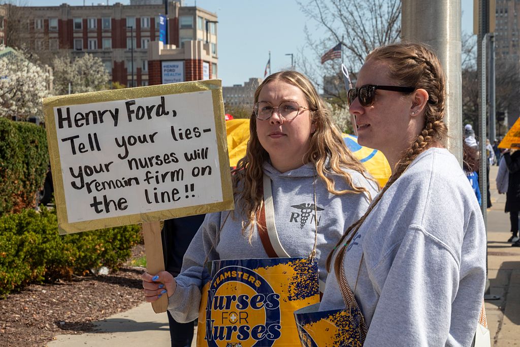 Two nurses on strike, one holds a sign that says, "Henry Ford, tell your lies—your nurses will remain firm on the line!"