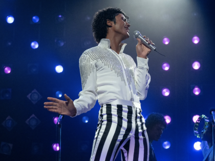 Jaafar Jackson as Michael Jackson in black-and-white-striped pants and a white shirt, singing into a microphone with a blue backdrop and lights.