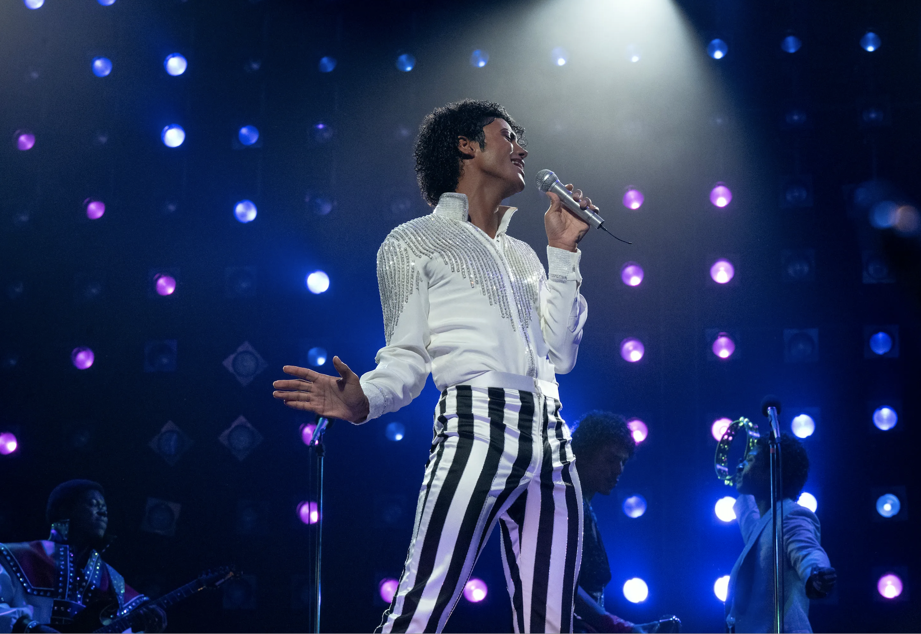 Jaafar Jackson as Michael Jackson in black-and-white-striped pants and a white shirt, singing into a microphone with a blue backdrop and lights.