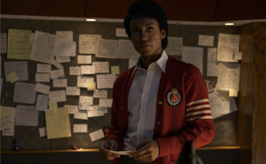 Actor Jaafar Jackson portrays Michael Jackson in a red cardigan with a bulletin board filled with paper behind him.