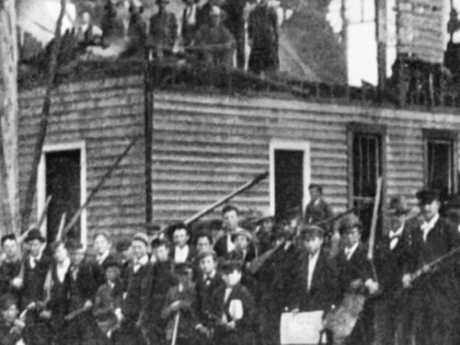 Armed militiamen stand in front and atop the burned building of the black newspaper, the Daily Record.