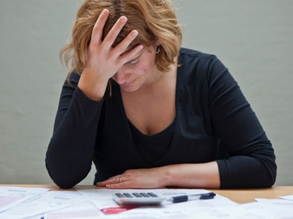 A visibly tired and distressed woman sits at a table with a stack of bills before her.
