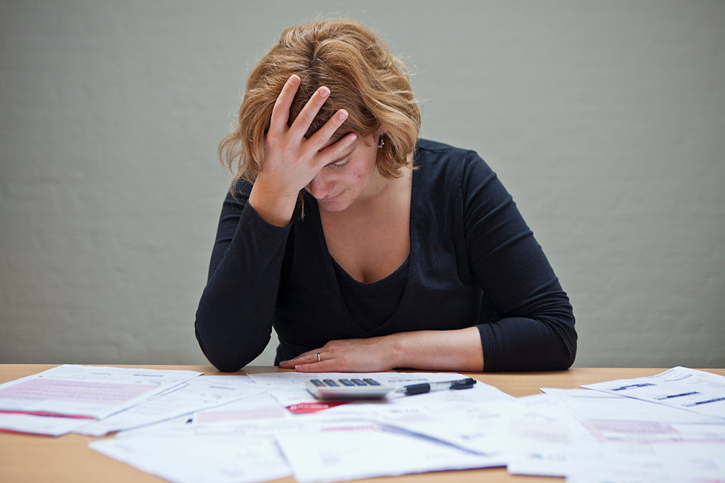 A visibly tired and distressed woman sits at a table with a stack of bills before her.