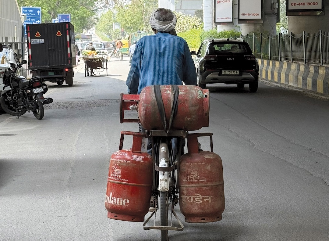 A man transports multiple LPG cylinders on a bicycle along a busy road in New Delhi, India.