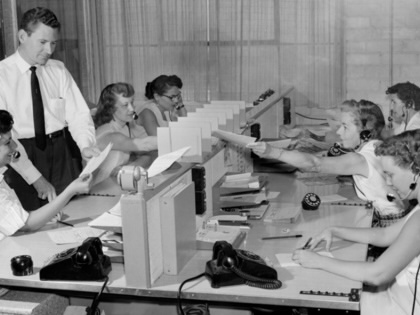 A vintage photograph of a team of women, seated, answering telephone calls as a man, standing, is shown some paperwork . 