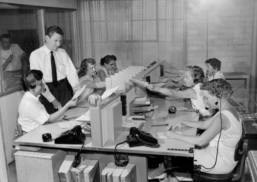 A vintage photograph of a team of women, seated, answering telephone calls as a man, standing, is shown some paperwork .